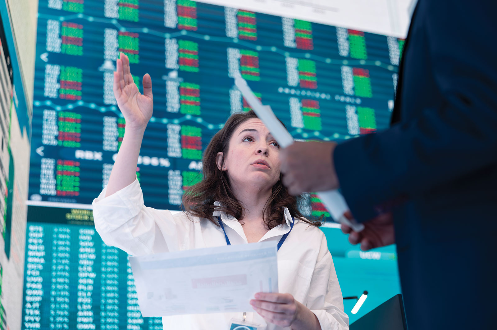 Woman raising her hand while reviewing documents in front of a stock market ticker display